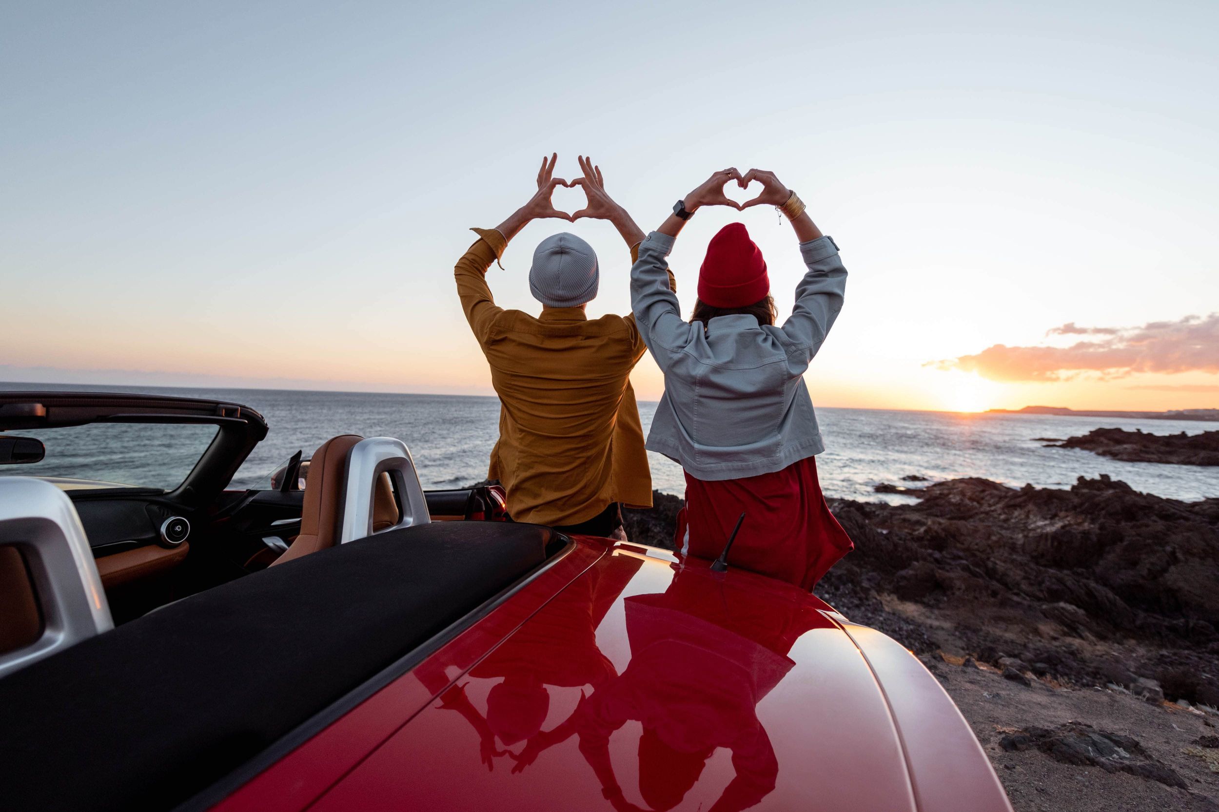 Zwei Jugendliche stehen gedreht zur Sonneuntergang vor einem roten Sportauto und zeigen mit beiden Händen ein Herzzeichen.